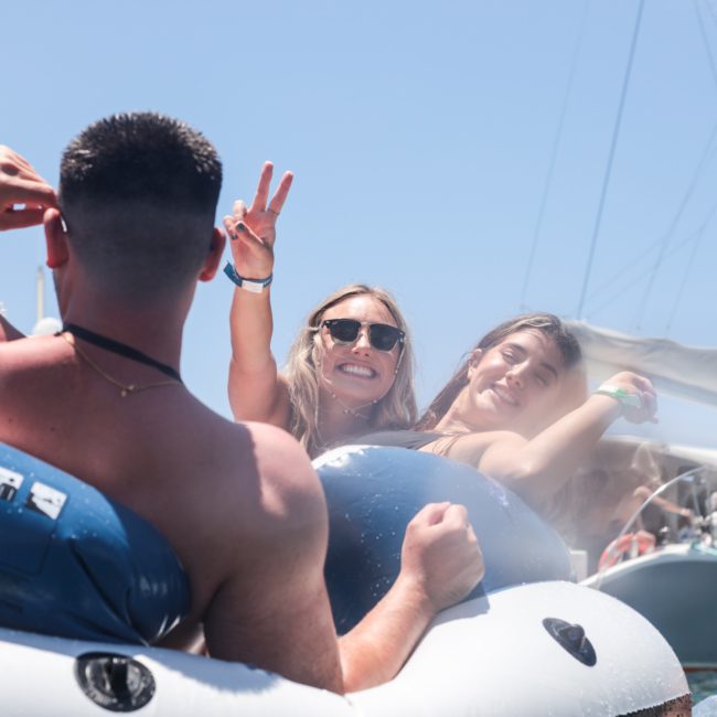 Three people are on an inflatable boat in the water, smiling and making hand gestures. The background shows a sailboat against a clear blue sky, perfect for a Sydney boat party hire.