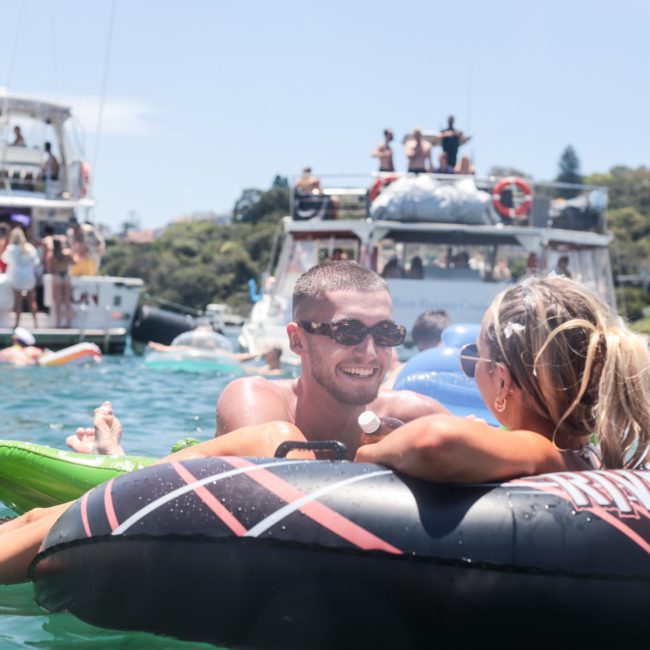 A man and a woman float on an inflatable tube in the water while talking. Nearby, other people enjoy themselves on boats and floating, taking in the serene surroundings. It’s a perfect day for a private yacht charter Sydney Harbour experience.