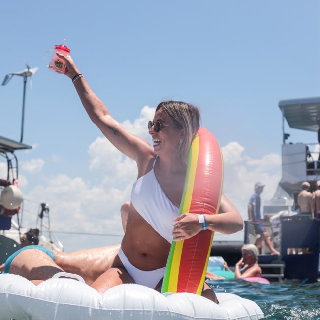 A woman in a white swimsuit is sitting on an inflatable pool float, holding a drink and cheering, with boats and other people enjoying the Luxury yacht hire Sydney experience in the background.