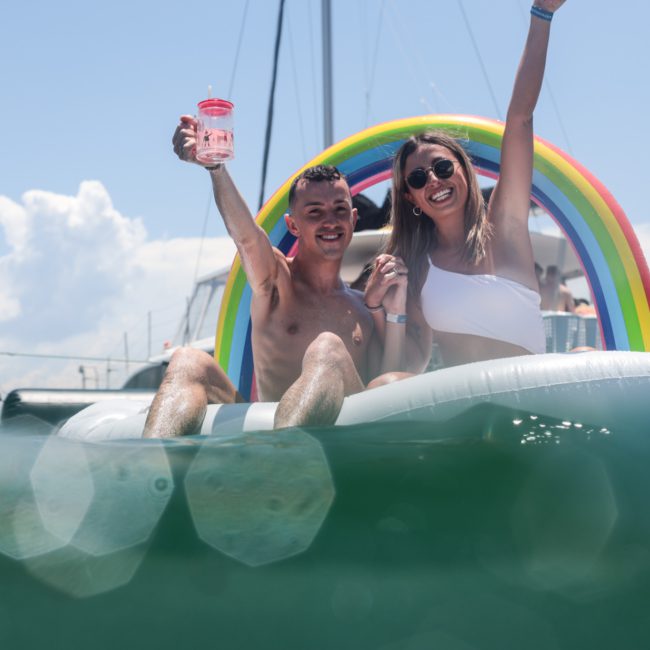 Two people sit on an inflatable rainbow float in the water, smiling and raising drinks. A private yacht charter in Sydney Harbour can be seen in the background under a clear sky.