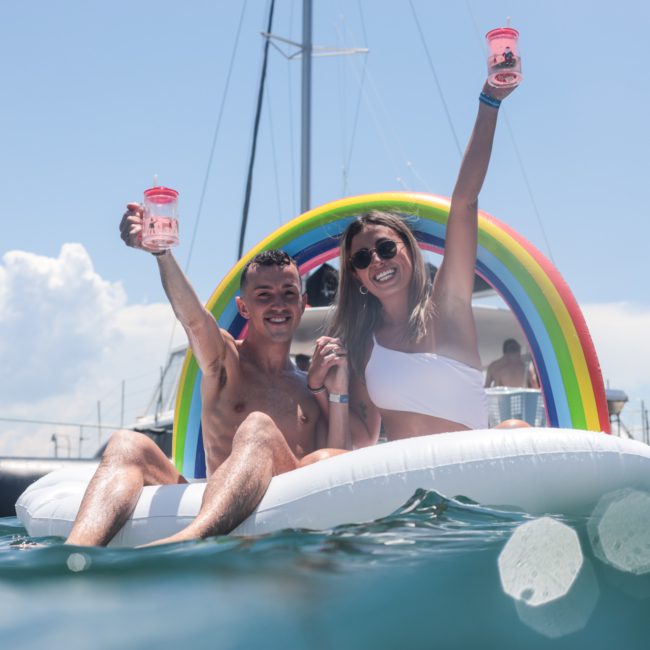 Two people sit on an inflatable float in the water, holding drinks and smiling, with a boat and rainbow arch in the background, showcasing the fun of a Sydney boat party hire.