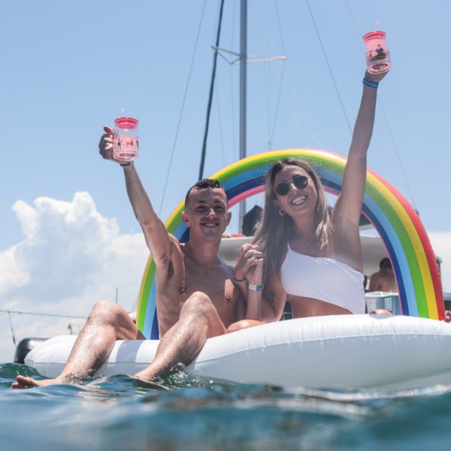 Two people smiling and raising cups while sitting on an inflatable boat with a rainbow arch, floating on water under a clear sky—perfect for a Corporate boat event in Sydney.