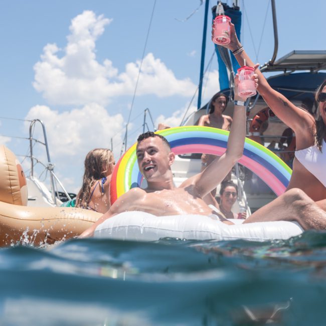 People relaxing on inflatable floaties in the water near a boat. A man and woman raise drinks in celebration while others enjoy themselves in the background. Sunny day with a few clouds in the sky, perfect for a private yacht charter on Sydney Harbour.