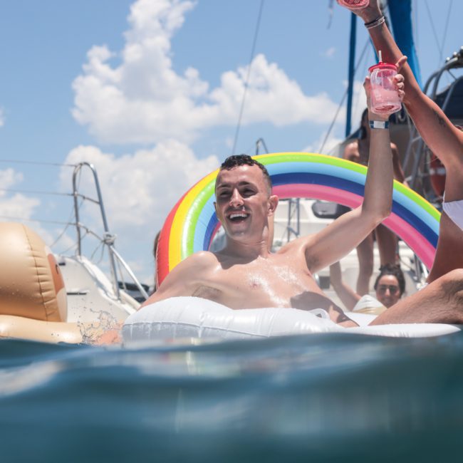 Two people celebrating on an inflatable float with a rainbow design in the water, near docked boats on a sunny day. Perfect for a private yacht charter Sydney Harbour experience!