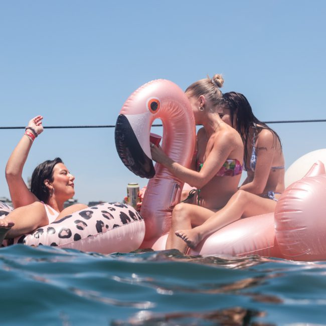 Three women are floating on inflatable pool toys in open water on a sunny day, enjoying a Sydney boat party hire. Two are on a flamingo floatie, and one is on a leopard-print floatie. One woman is raising her hand, smiling.