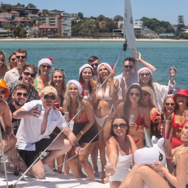 A group of people in swimsuits and festive hats are gathered on a sailboat, smiling and holding drinks. The backdrop includes a coastal town and clear blue water under a sunny sky, perfect for luxury yacht hire Sydney.