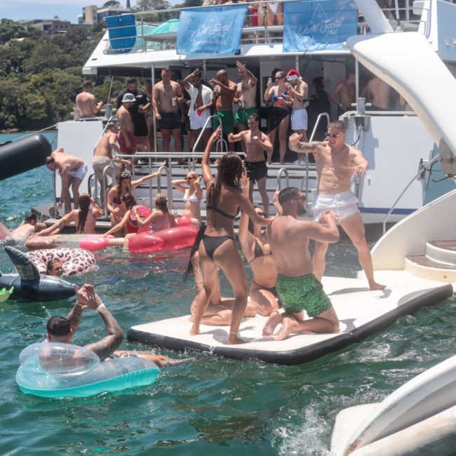A group of people enjoy a sunny day on a private yacht charter in Sydney Harbour, swimming, lounging on inflatable floats, and socializing in swimwear. Some stand on a floating platform by the boat's deck.