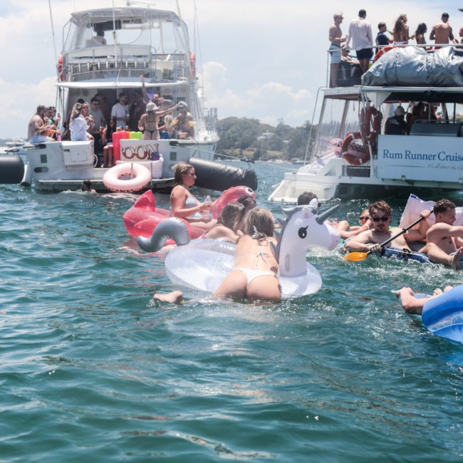 A group of people in swimsuits are relaxing on inflatable floats in the water near anchored boats under a sunny sky, enjoying a fantastic Sydney boat party hire.