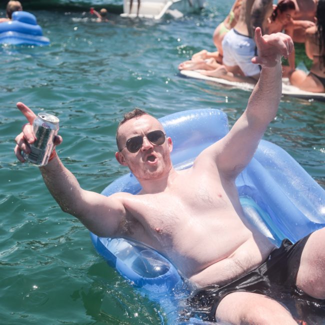 A man in sunglasses and swim shorts holds a can and gestures energetically while floating on a blue inflatable raft in a body of water with other people socializing in the background, enjoying a catamaran party Sydney style.