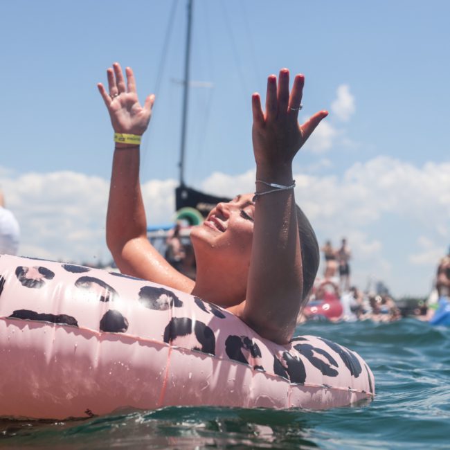 Person in a leopard-print inflatable inner tube enjoying the water with arms raised, surrounded by other swimmers and boats on a sunny day. Nearby, people revel in the fun atmosphere of a luxury yacht hire Sydney, creating perfect summer memories.