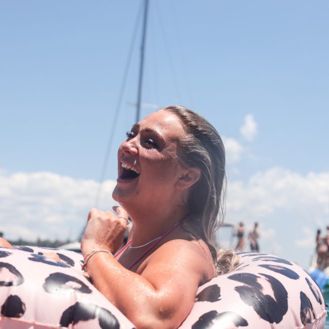 A person smiling while floating on an inflatable tube with a leopard print design in a body of water, with luxury yachts and other people enjoying the scene in the background.