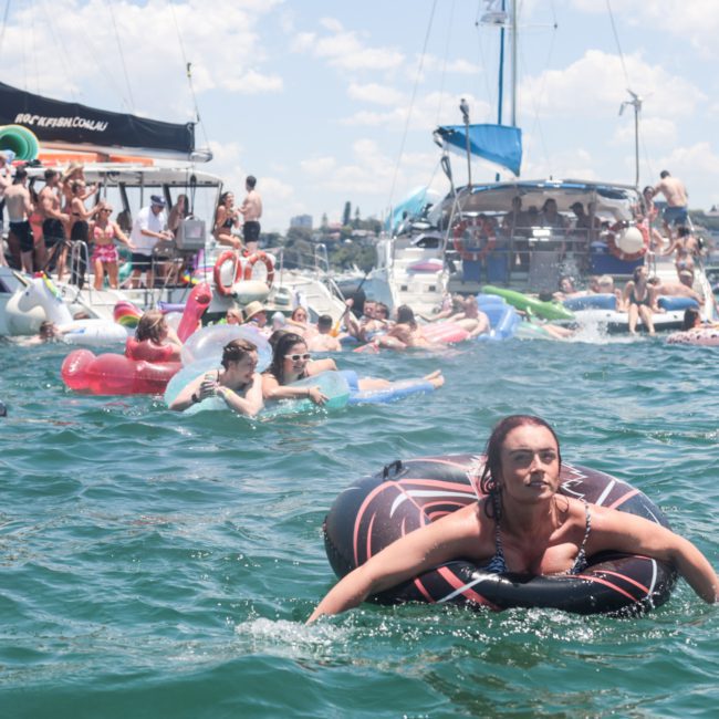 A crowded scene of people on boats and floating on inflatables in a sunny day on the water, with one woman in the foreground on a black inflatable ring. In the distance, luxurious catamaran party Sydney vibes can be seen.