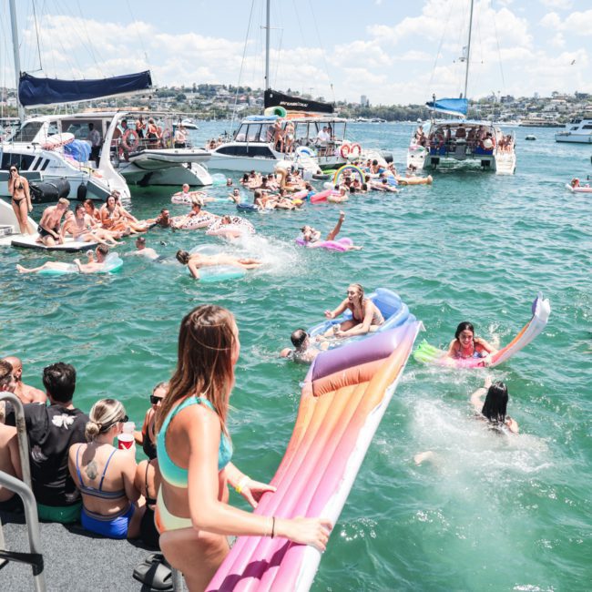 A large group of people in swimwear enjoys a daytime boat party on a lake, with some on boats and others swimming or floating on inflatables in the water. It's reminiscent of a lively Catamaran party in Sydney, perfect for corporate boat events.