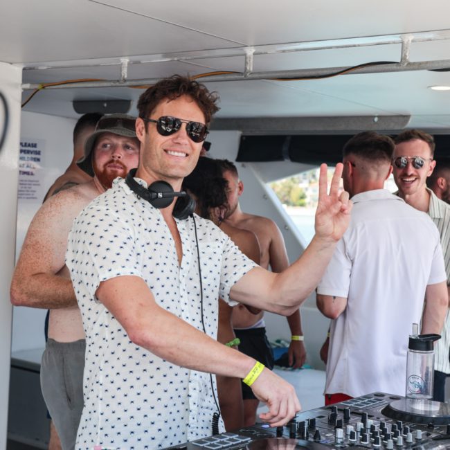 A man with headphones and sunglasses stands in front of DJ equipment, flashing a peace sign. Behind him, a group in swimwear enjoys the scene on a luxury yacht hire in Sydney.