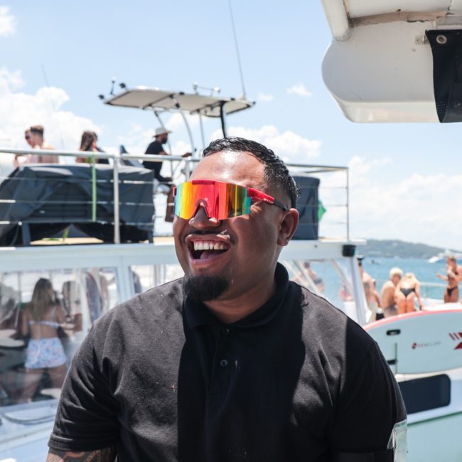 A person wearing colorful sunglasses smiles on a catamaran party in Sydney Harbour with a crowd of people and another boat in the background on a sunny day.