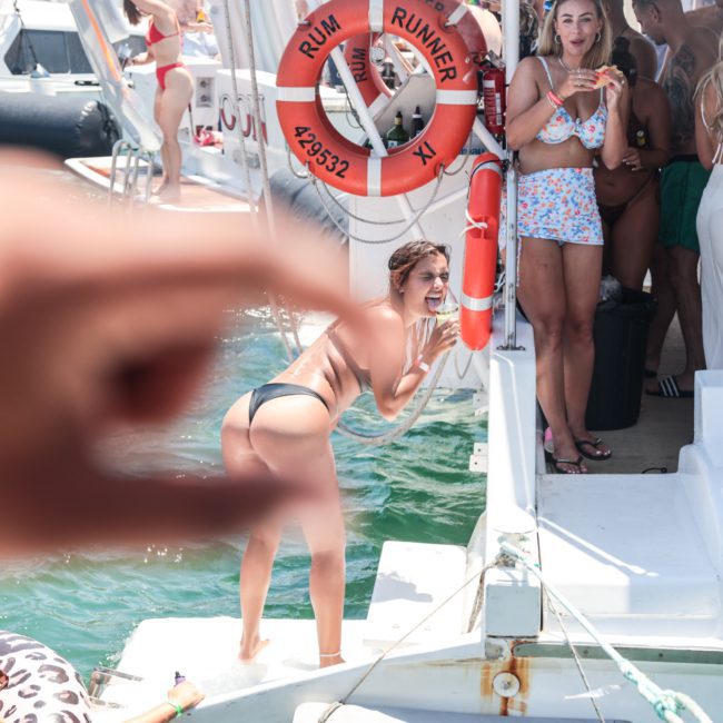 A woman in a bikini poses near a boat while a hand in the foreground makes a pinching gesture, creating a playful forced perspective. Other people are gathered on the boat in the background, enjoying what looks like an amazing corporate boat event in Sydney.