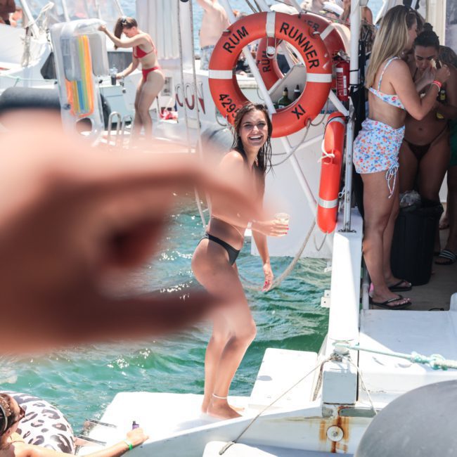 A woman in a black bikini poses and laughs on a boat dock as a hand in the foreground appears to pinch her. Other individuals are seen enjoying a luxury yacht hire on Sydney Harbour in the background.