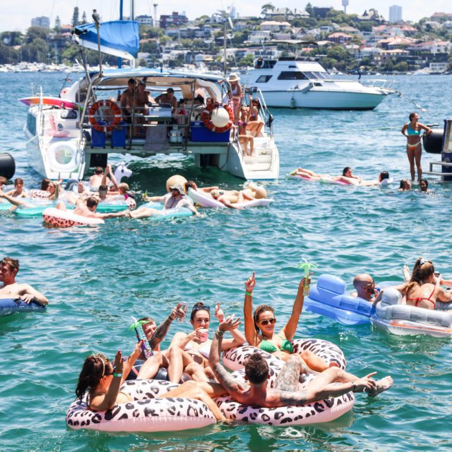 People lounge on inflatable floats in the water near boats, enjoying a sunny day at a lively waterfront gathering, reminiscent of private yacht charters in Sydney Harbour.