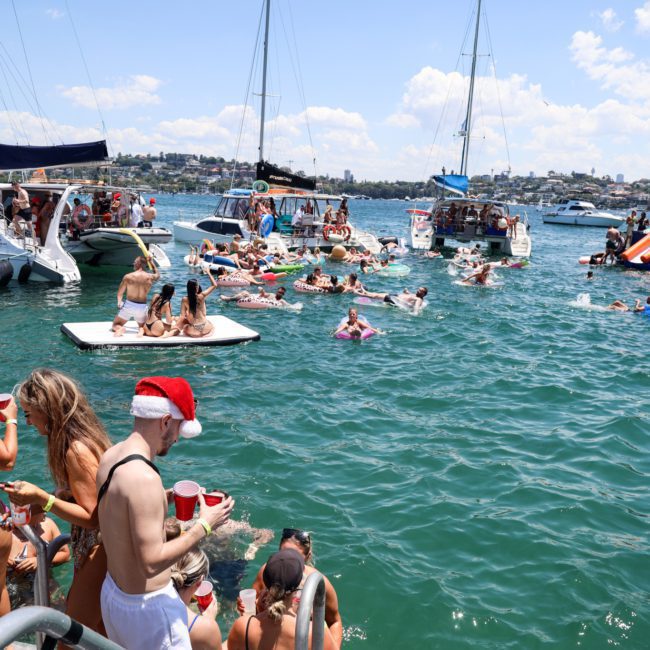 A crowded boat party on a sunny day with people in swimsuits swimming, floating, and socializing; some wear Santa hats and hold red cups. Various boats, including those from private yacht charter Sydney Harbour, and a large luxury yacht are docked nearby.
