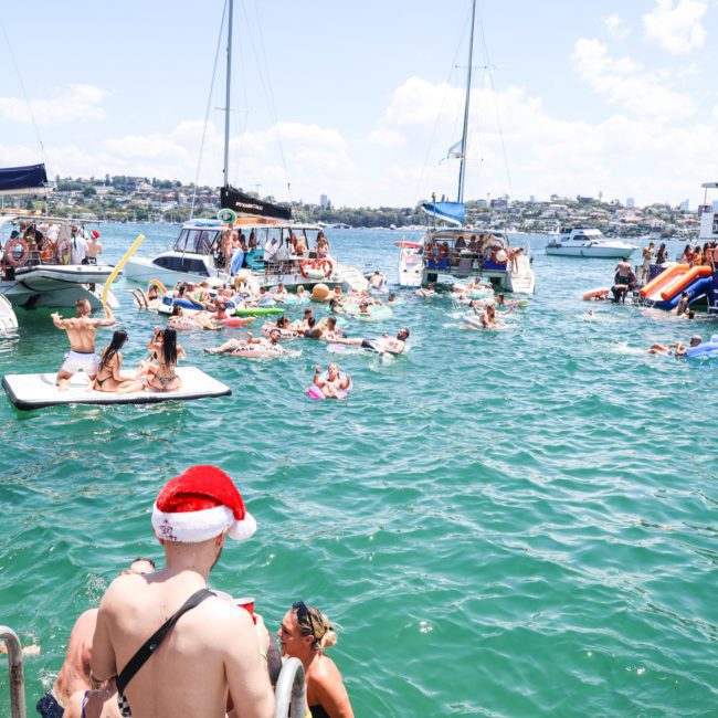 People enjoying a sunny day on the water, lounging on floats and boats with a city skyline in the background. One person wears a Santa hat during their luxury yacht hire in Sydney.