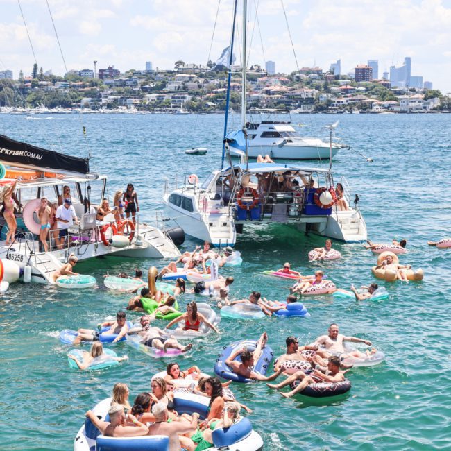 Groups of people on boats and inflatables enjoy a sunny day on the water near a coastal town, some indulging in luxury yacht hire Sydney for an exclusive experience.