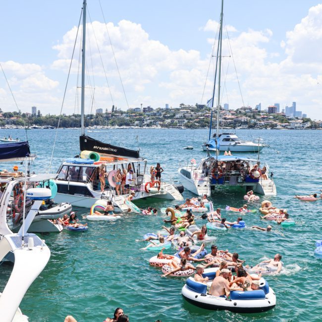 People relax and socialize on a catamaran party in a sunny harbor with a cityscape in the background.