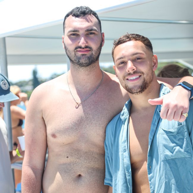 Two individuals pose for a photo outdoors, one shirtless and wearing blue shorts, the other in a blue shirt with the top buttons undone. They are under an awning with people and the sky in the background, hinting at a fun Sydney boat party hire event.