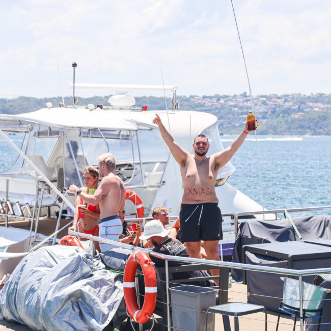 A person stands on the deck of a catamaran party in Sydney, holding drinks and raising their arms. The lively crowd surrounds them, as they enjoy the scenic views of Sydney Harbour's distant shoreline in the background.