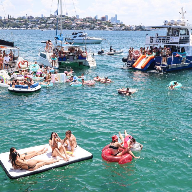 Groups of people relax on floating platforms and inflatable devices in a vibrant bay with several anchored boats and yachts nearby, ideal for a lively Sydney boat party hire.