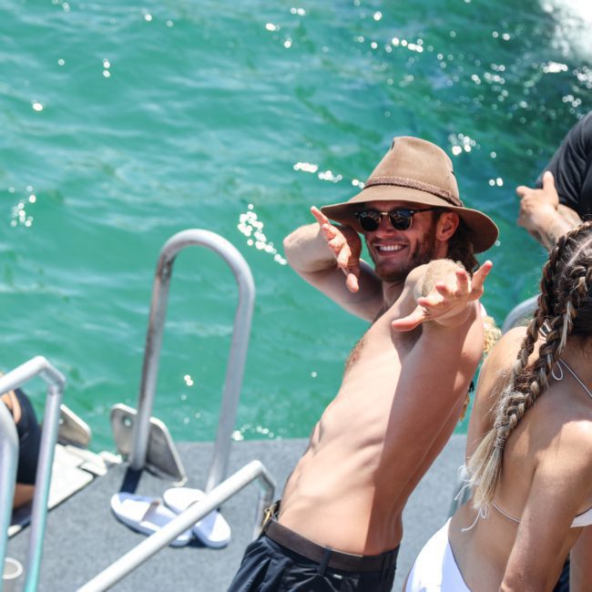 A man in a sunhat poses and smiles on a private yacht charter in Sydney Harbour with others near the water. Several people are standing nearby, some holding red cups.