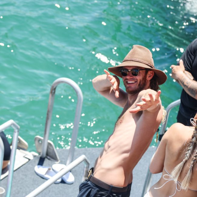 A man wearing a hat and sunglasses poses playfully on a boat surrounded by other people, with the sparkling waters in the background—a perfect scene for a private yacht charter Sydney Harbour.