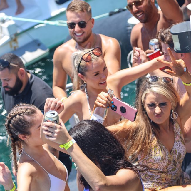 A group of people in swimwear are partying on a private yacht charter in Sydney Harbour, holding drinks and taking selfies. The water and another boat are visible in the background.