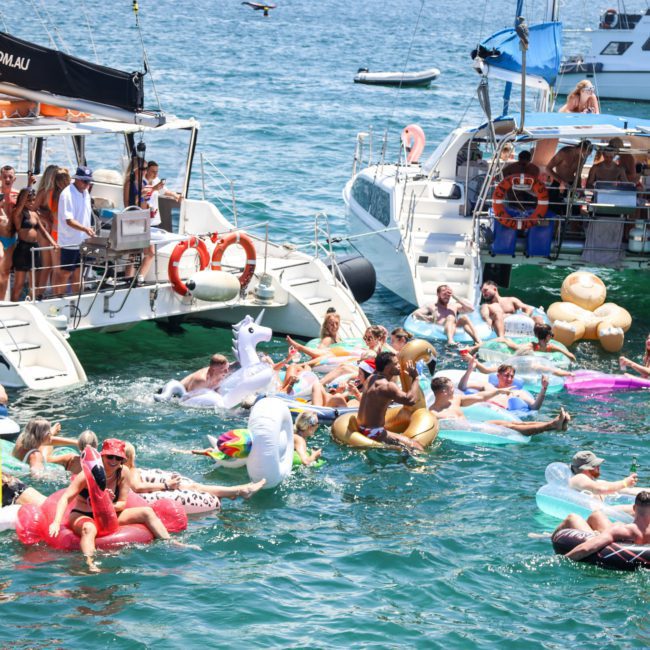 People floating on inflatable pool toys in the water near two boats. The scene appears lively with a mix of colorful inflatables and calm blue water, perfect for a private yacht charter in Sydney Harbour.