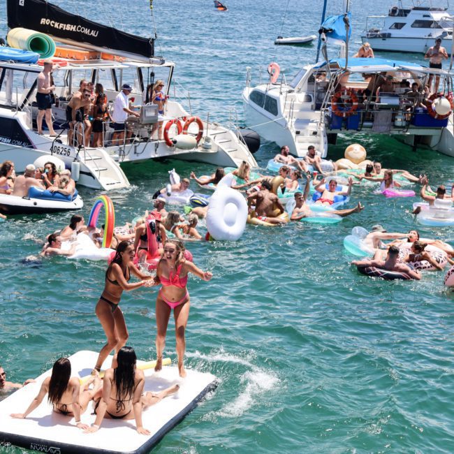 A group of people enjoying a sunny day on boats and inflatable floats in the water, with some indulging in a luxurious Catamaran party in Sydney. Others relax on various inflatable toys, making the most of the beautiful weather.