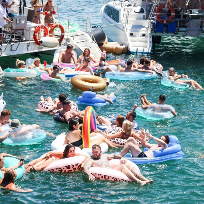 People enjoying a sunny day on colorful inflatable floats in the water near several boats. The scene is lively with groups of friends gathered together, highlighting the perfect ambiance for a private yacht charter in Sydney Harbour.
