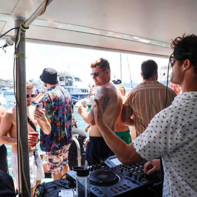 A DJ playing music on a boat deck while a group of people socialize and enjoy the sunny weather on a private yacht charter in Sydney Harbour, with other boats visible in the background on the water.