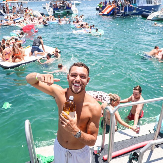 A man stands on a boat holding a large bottle, smiling at the camera. In the background, numerous people are swimming, floating on inflatables, and socializing near several anchored boats. It's the perfect scene for a Catamaran party in Sydney.