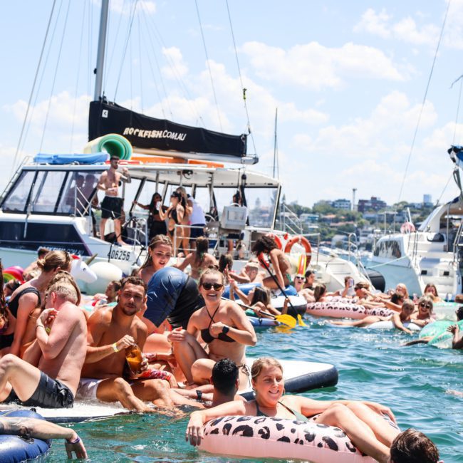 A crowd of people on inflatable floats and boats enjoying a sunny day on the water. The scene includes various types of boats, including some participating in luxury yacht hire Sydney, with individuals swimming and lounging in the water.