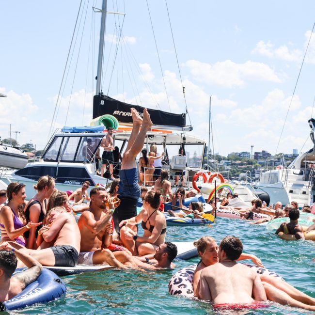 A group of people on inflatable floats enjoys a sunny day in the water, surrounded by moored boats, as the DJ boat hire Sydney service sets the perfect vibe.