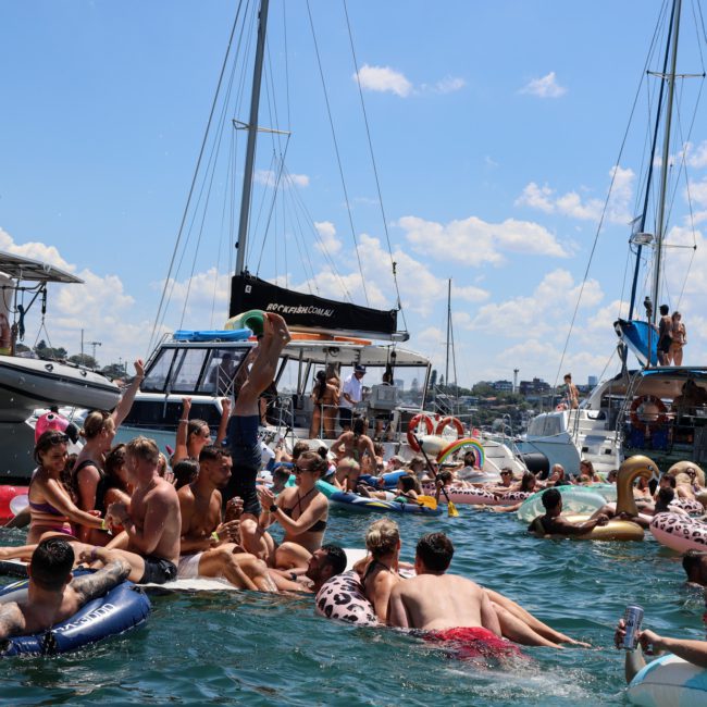 People on boats and inflatables are gathered together in a lively, crowded scene on the water under a partly cloudy sky, enjoying a catamaran party Sydney.