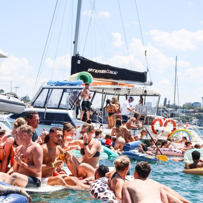 A large group of people enjoys a sunny day on the water, swimming and floating on inflatables near several moored boats. Many are celebrating with a Sydney boat party hire, complete with music and good vibes under the sun.