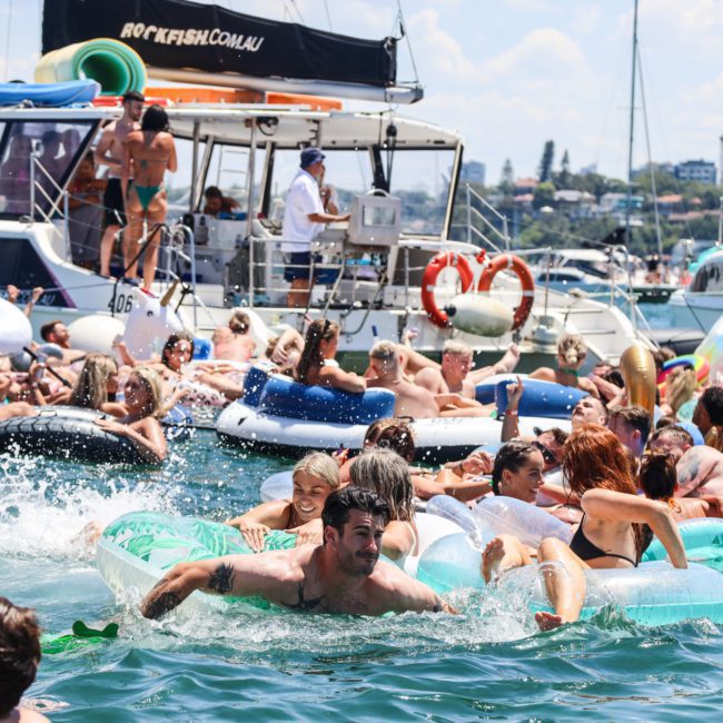 A large group of people enjoy the water on inflatables near docked boats on a sunny day. Some are swimming while others relax on large floaties, all part of a Sydney boat party hire event.