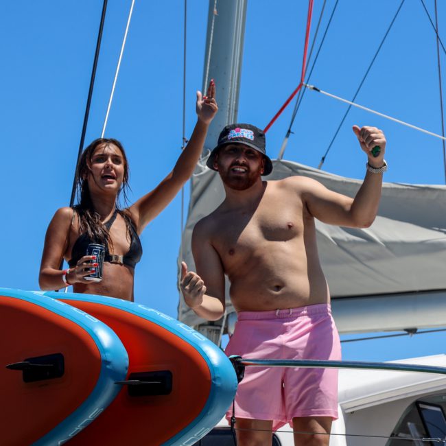 Two people stand on a boat deck; a woman in a bikini holds a drink and raises her hand, while a man in pink shorts and a bucket hat gestures with both hands, enjoying a luxury yacht hire Sydney experience.