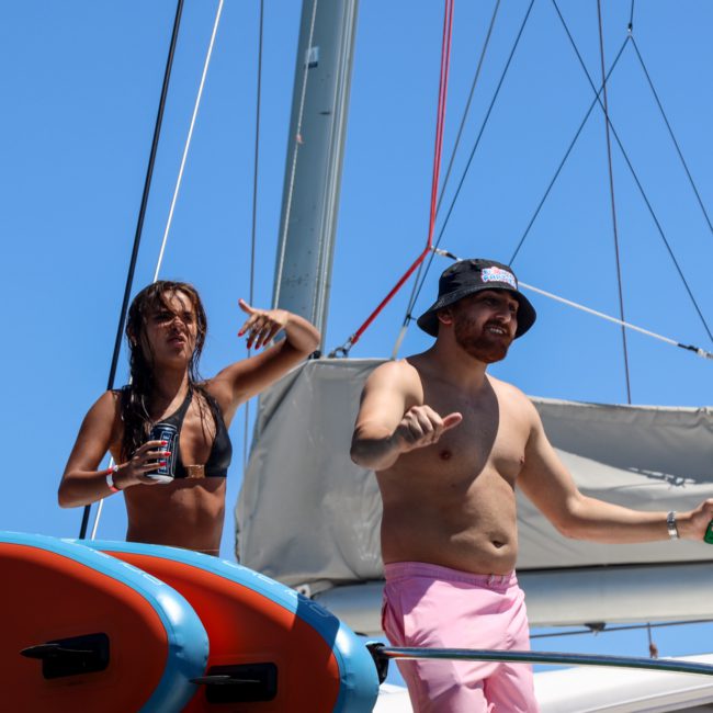 Two people in swimwear stand on the deck of a sailboat, one holding a drink and gesturing energetically while the other stands beside them in pink shorts and a black hat. Paddleboards are in the foreground, showcasing an ideal Corporate boat events Sydney experience.