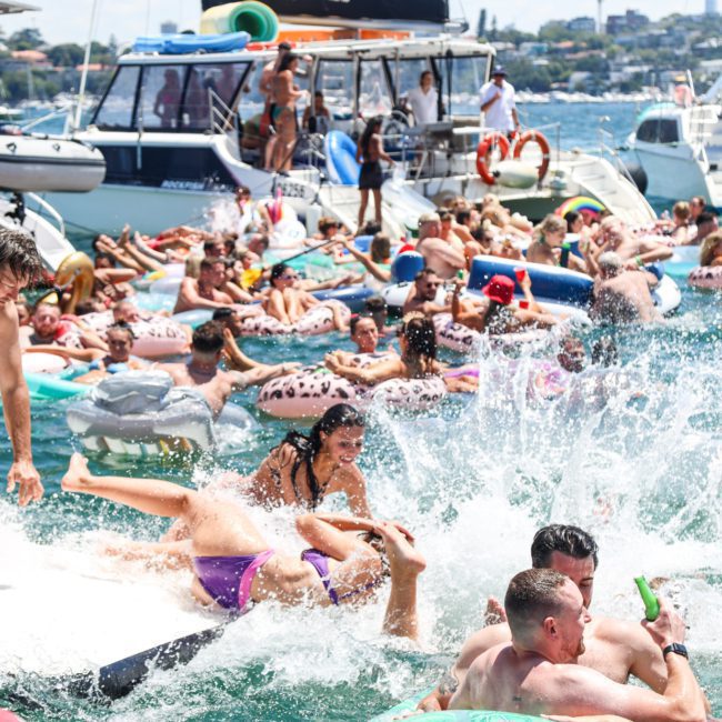 A crowded scene of people enjoying a summer day on the water, with some floating on inflatables and others on boats, creating a lively, festive atmosphere. The highlight is a luxury yacht hire Sydney experience that adds an extra touch of elegance to the celebration.