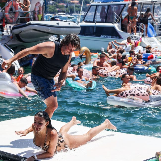 A man in a black tank top and blue shorts helps a woman in a leopard print swimsuit on a floating platform among a lively group of people in the water, with boats and luxurious yacht hire Sydney in the background.