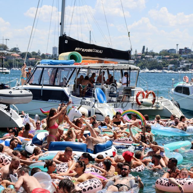 A crowd of people in swimsuits enjoying a private yacht charter Sydney Harbour with inflatable floats on a sunny day, surrounded by several boats on a body of water.