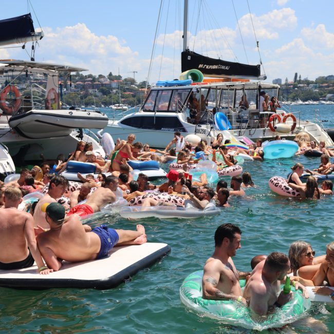 A group of people are enjoying a sunny day, floating on various inflatables and swimming in the water near several docked boats, making the most of their Sydney boat party hire.