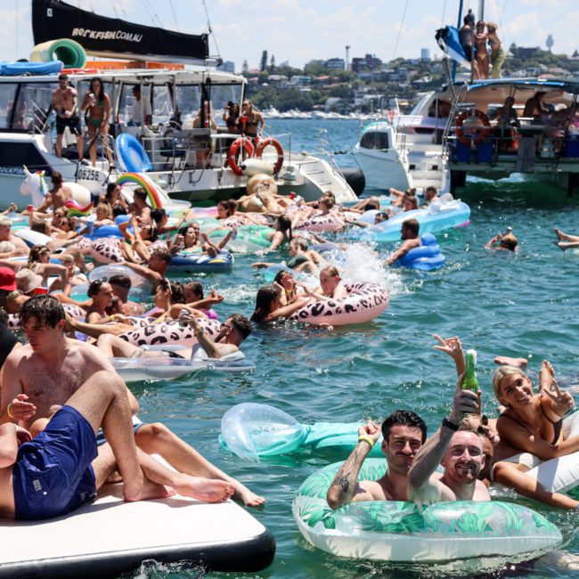 A crowded party with people on inflatable floats and boats in a sunny, waterfront setting, reminiscent of a Sydney boat party hire event.