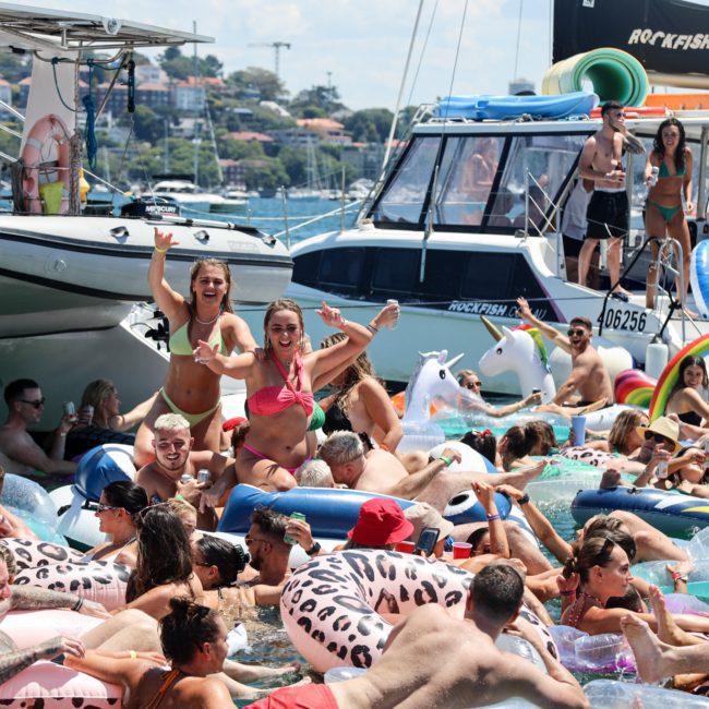 A crowded group of people enjoy a sunny day on inflatable floats in the water between boats. Some are waving and standing, while others relax on large pool floats during a fun-filled Catamaran party Sydney.